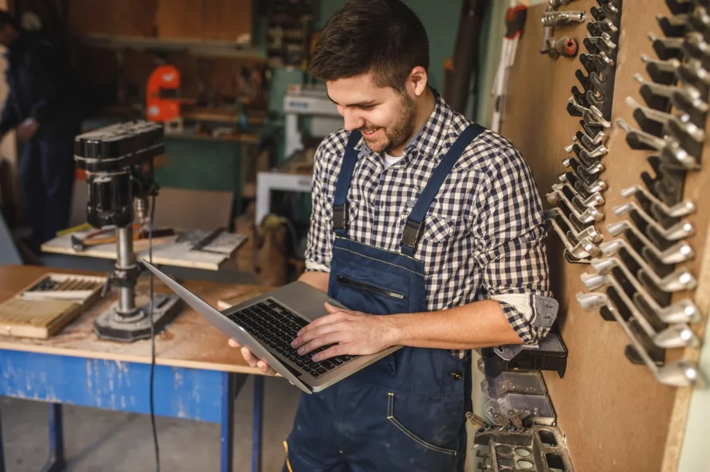 Handwerker schaut in seiner Werkstatt lächelnd auf den Laptop